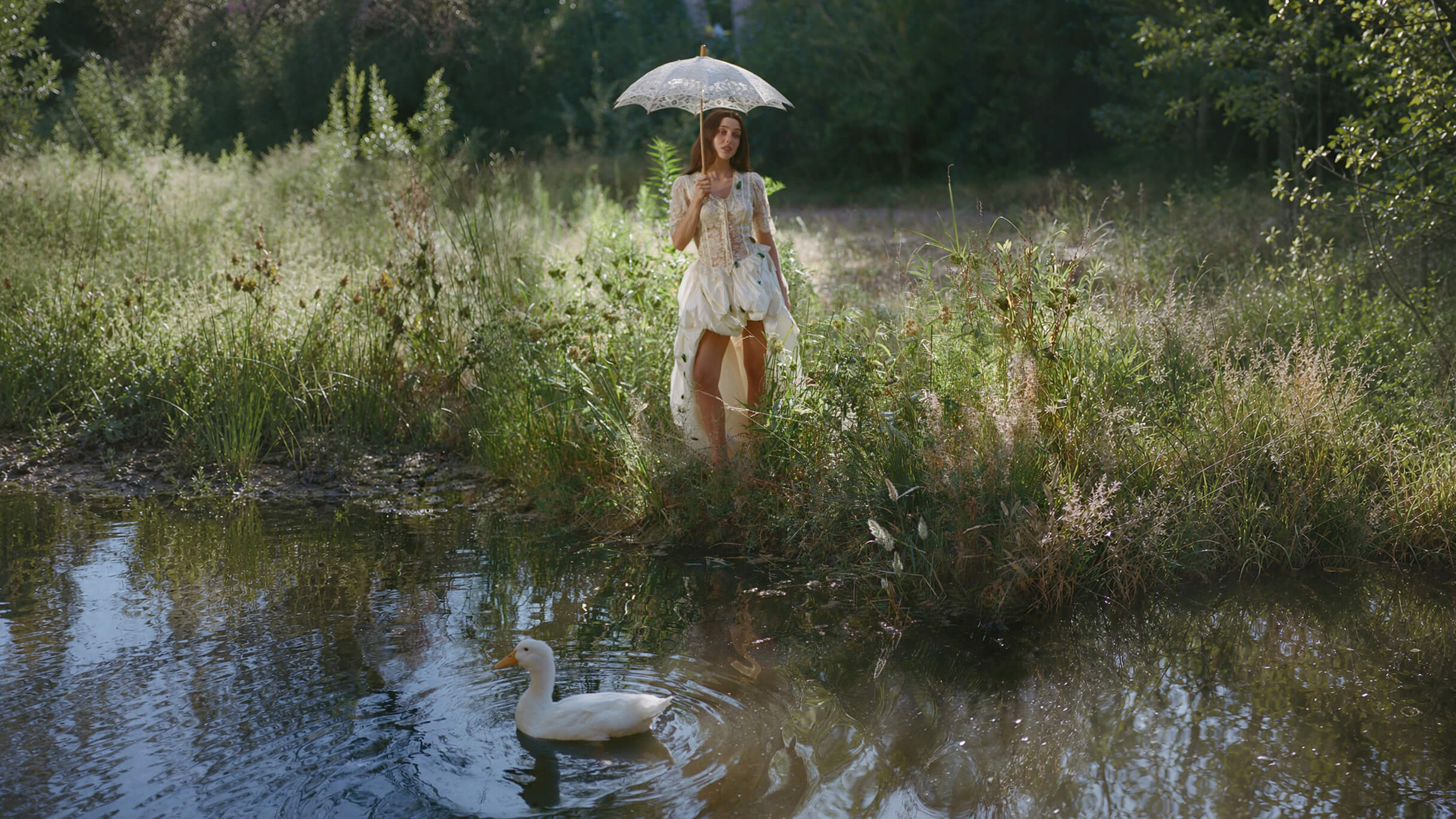 A woman in a flowing white dress stands beside a sunlit pond holding a lace parasol, while a white duck swims in the water below.