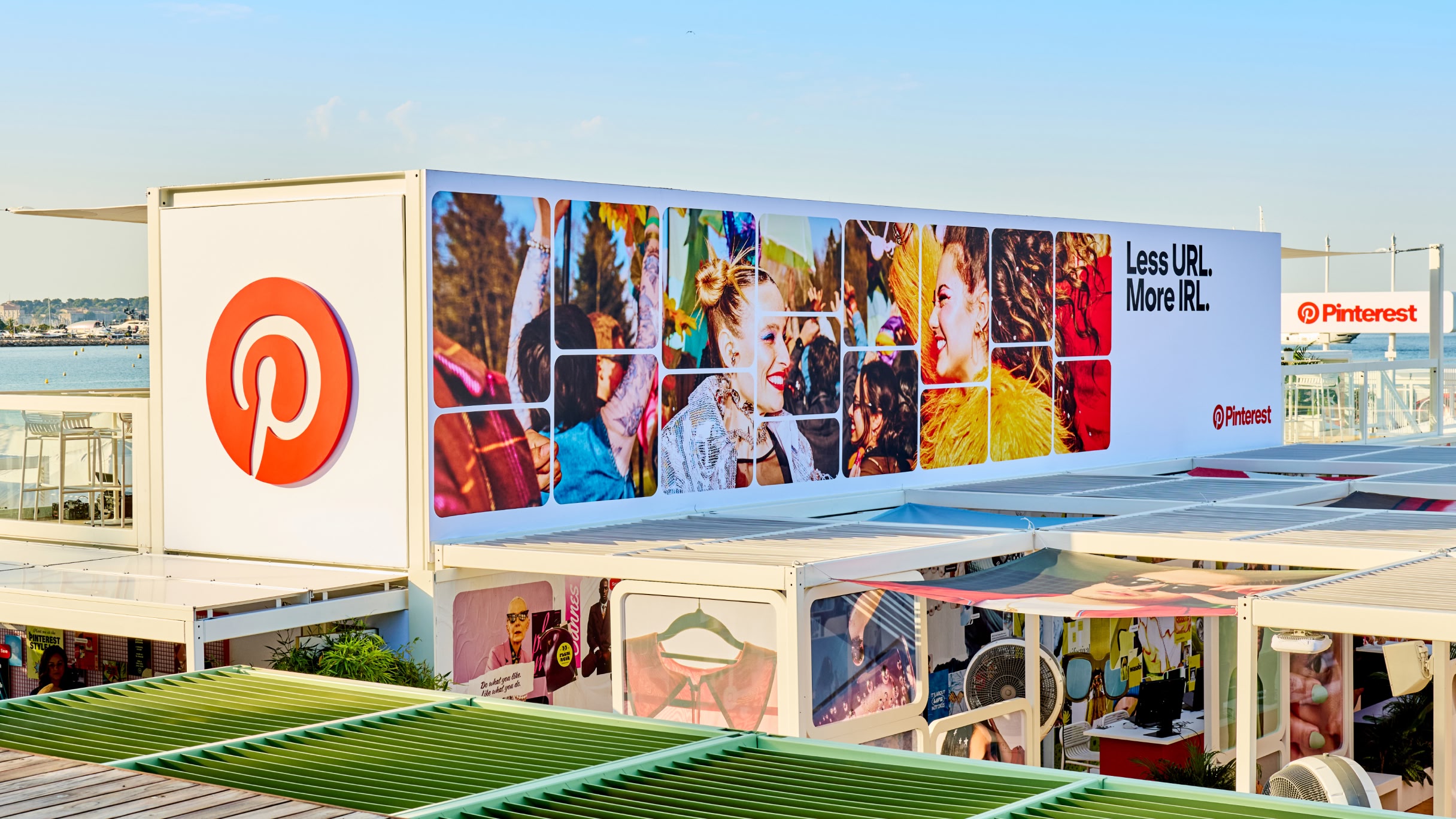 Vista externa do espaço Manifestival do Pinterest em Cannes, com um grande logótipo do Pinterest e um painel com fotos coloridas e o slogan “Less URL. More IRL.” diante de um cenário à beira-mar.