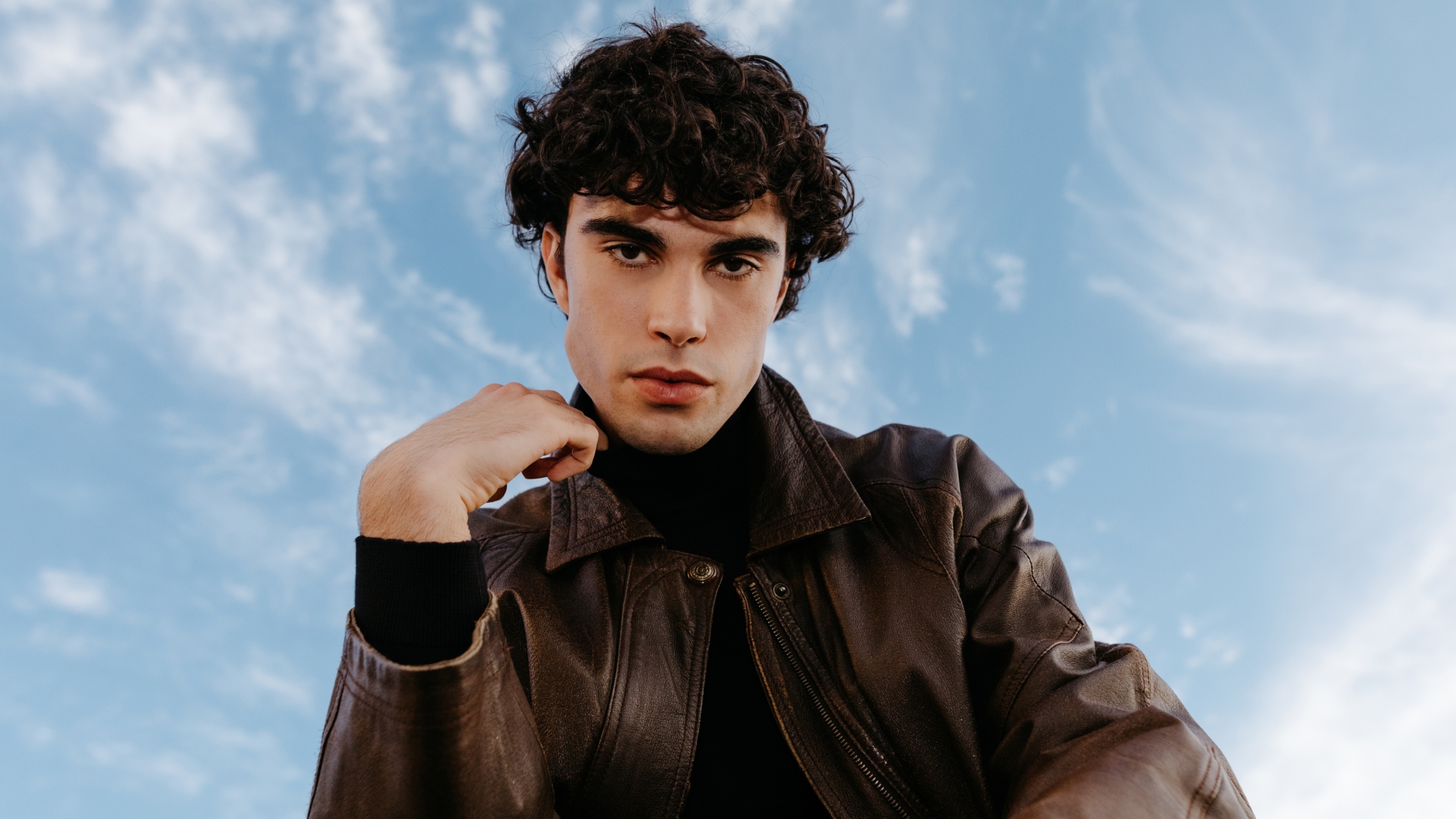 A person in a dark brown leather jacket and black turtleneck poses against a bright blue, cloud-filled sky.