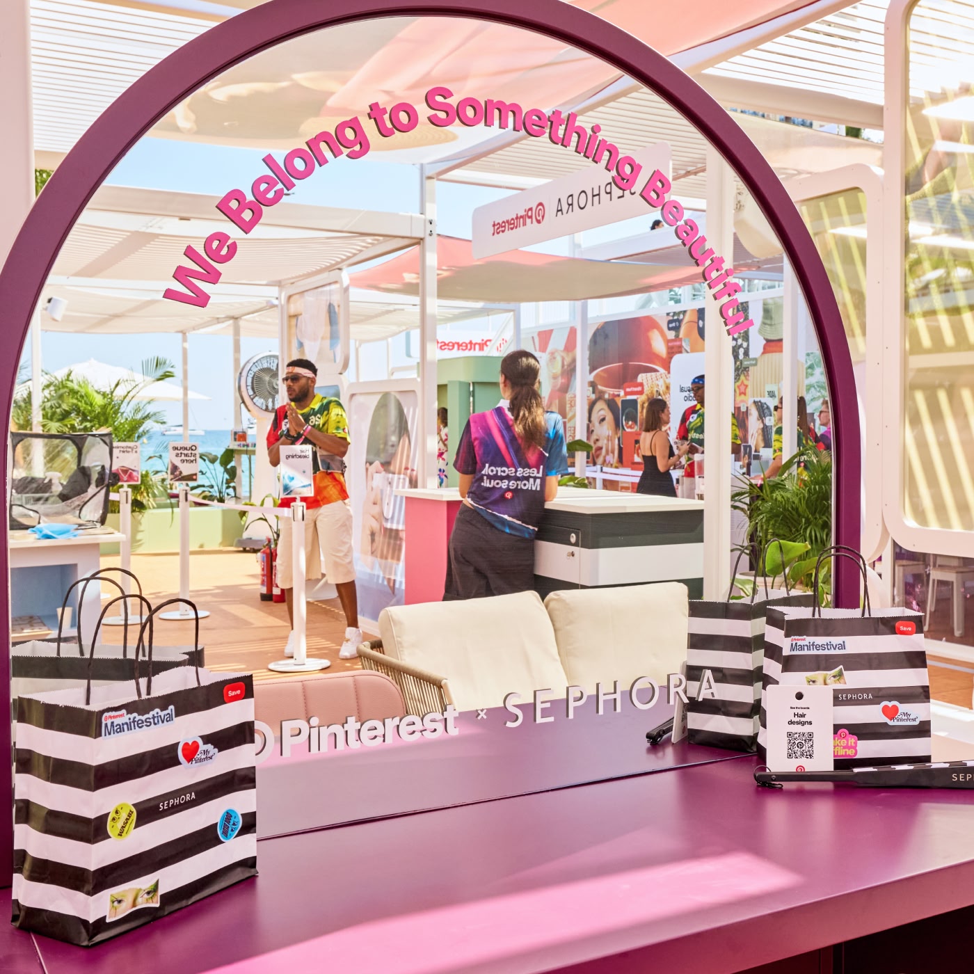 Una estación de belleza de Pinterest x Sephora en Pinterest Manifestival muestra un tocador con espejo morado con la frase “We Belong to Something Beautiful”, bolsas de marca y visitantes al fondo.