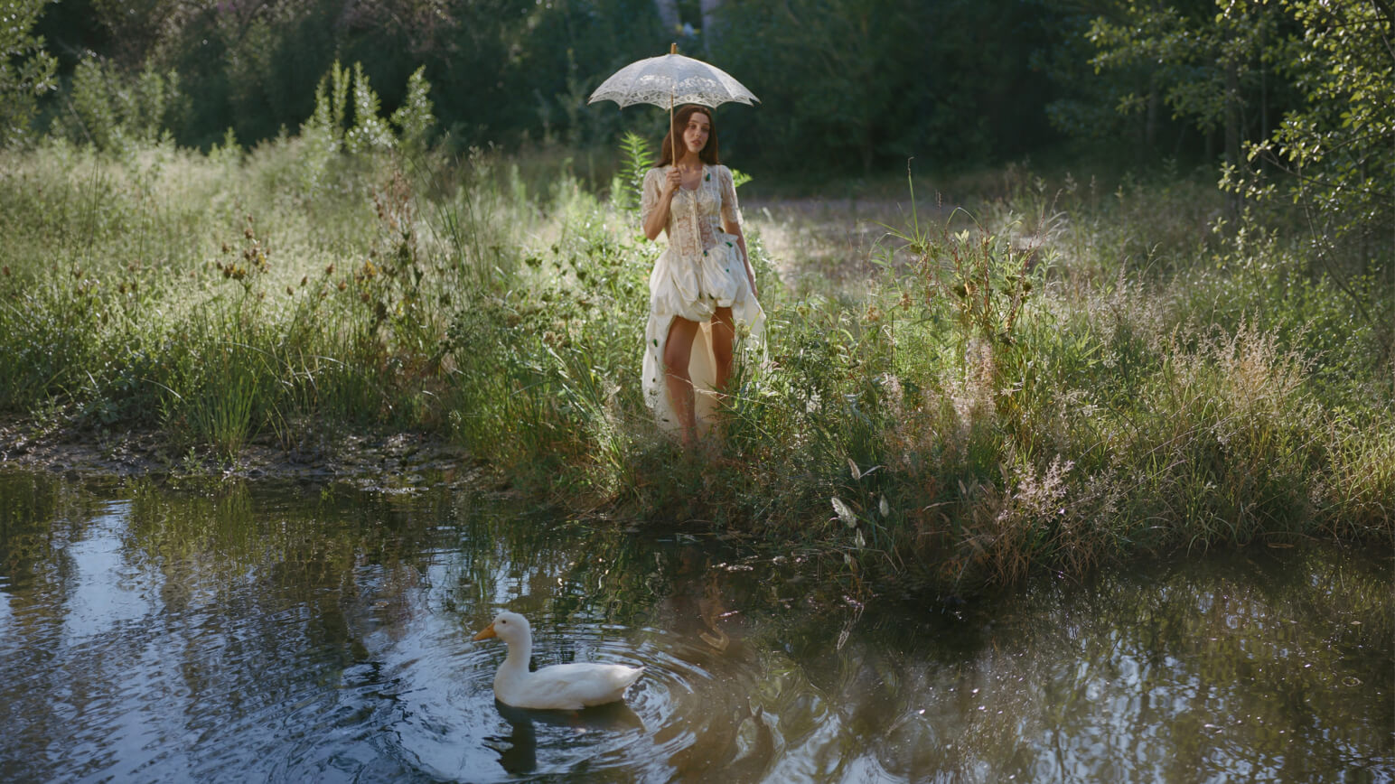 A woman in a flowing white dress stands beside a sunlit pond holding a lace parasol, while a white duck swims in the water below.