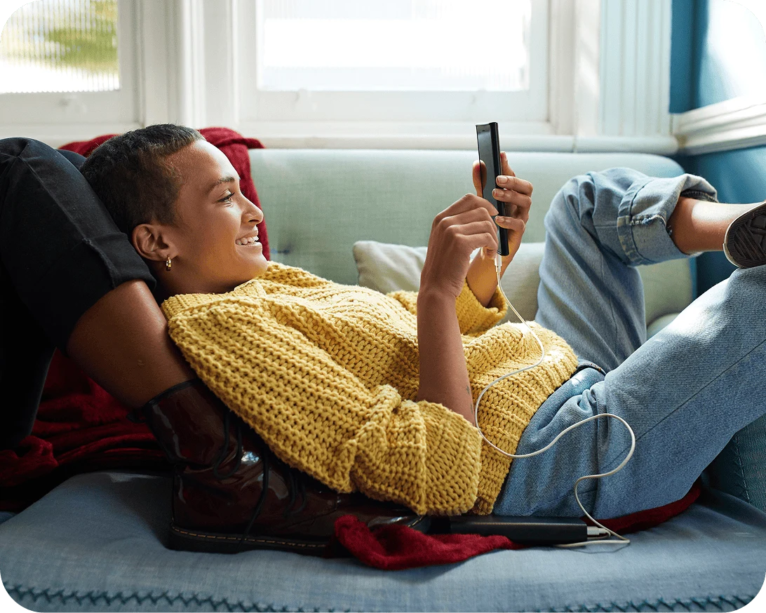 A woman with a yellow sweater lounges on  a couch while smiling and looking at her phone.