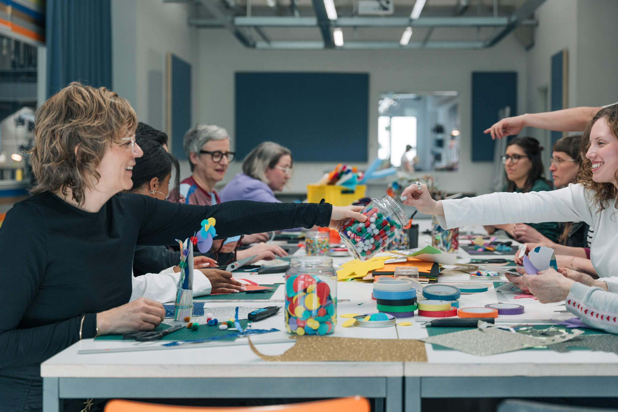 Eine Gruppe von Menschen sitzt an einem langen Tisch bei einem Kreativ-Workshop. Sie teilen Bastelmaterialien wie Washi-Tape, buntes Papier und Pompons und arbeiten lachend zusammen.