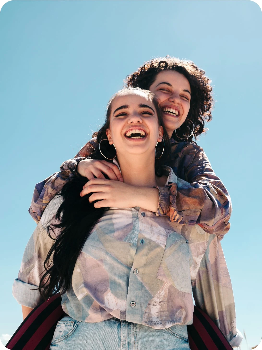 Two friends are laughing and hugging against a pale blue sky