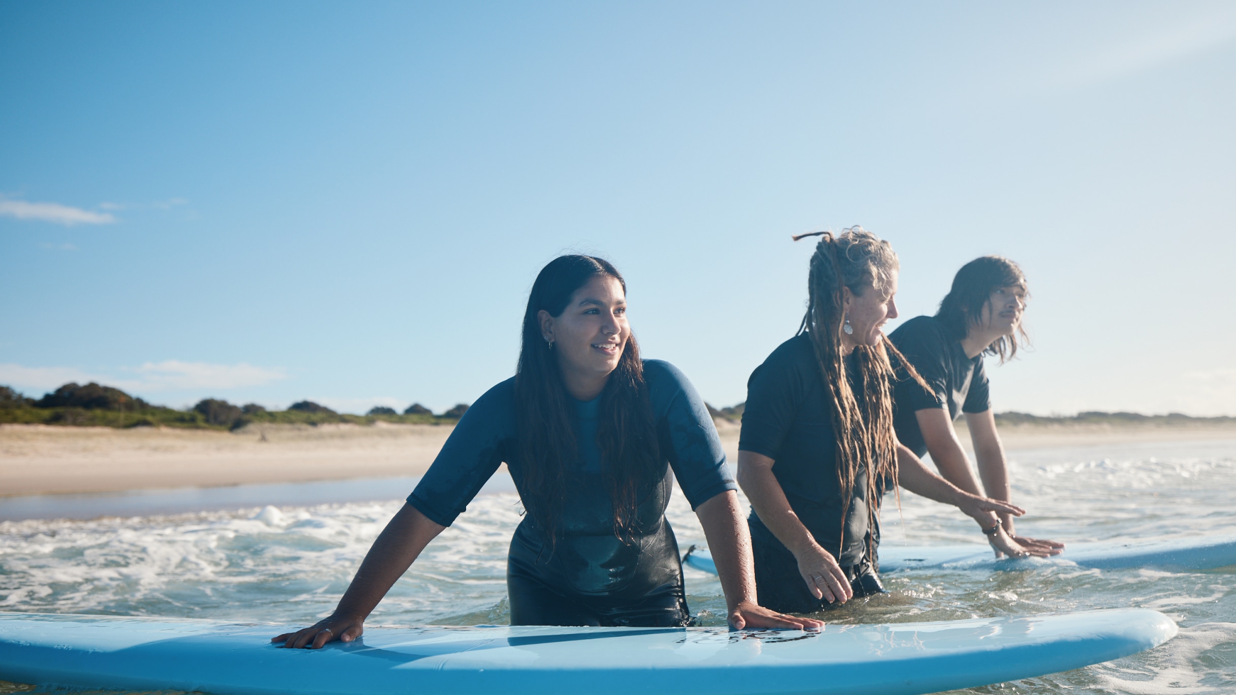 Three people in wetsuits standing in shallow ocean water, smiling and holding onto surfboards under a clear blue sky.