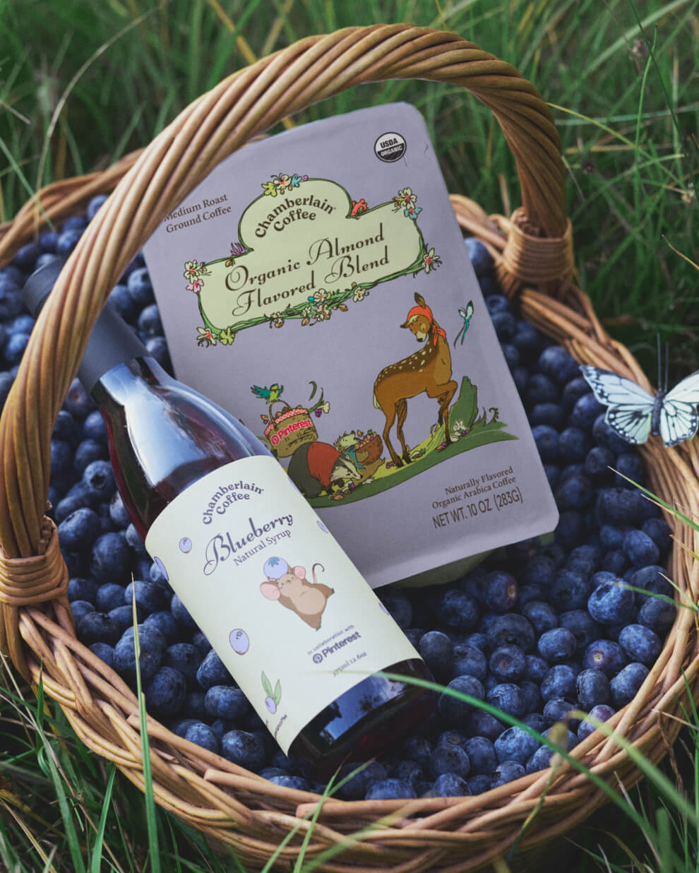 A basket of blueberries holding Chamberlain Coffee’s almond blend and blueberry syrup in tall grass, with a butterfly nearby.