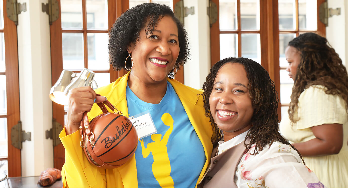Two women pose smiling at the camera with one holding a purse designed like a basketball. 
