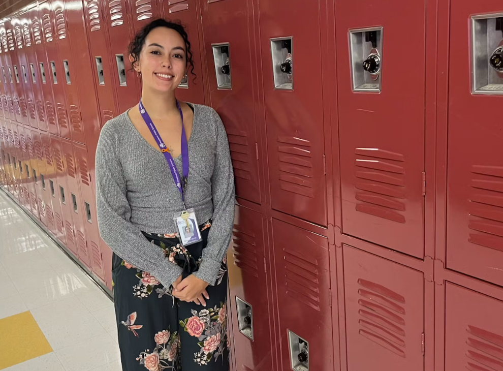 Teacher smiling while standing in a hallway and leaning against a row of red school lockers.