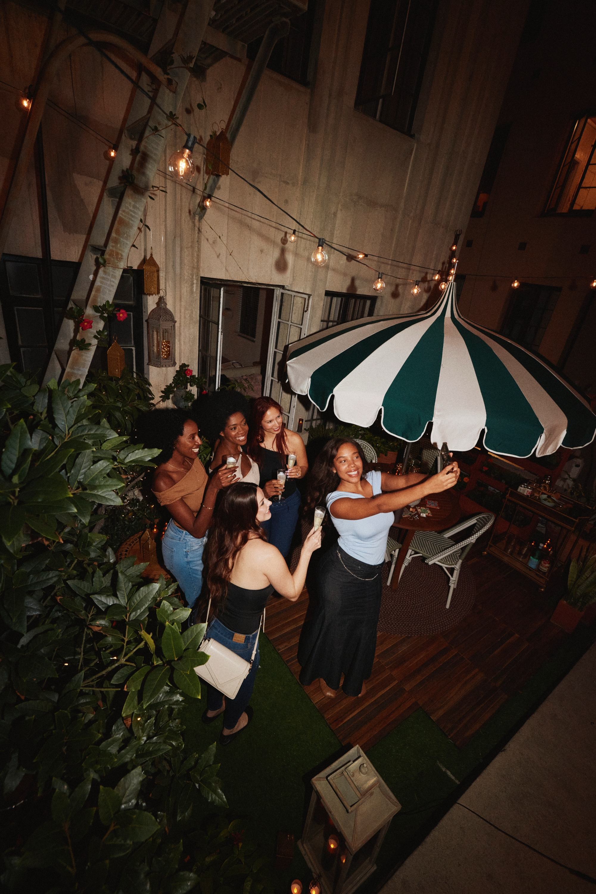 Five friends on a cozy outdoor patio at night with one woman taking a group selfie under string lights and a green-and-white striped umbrella.