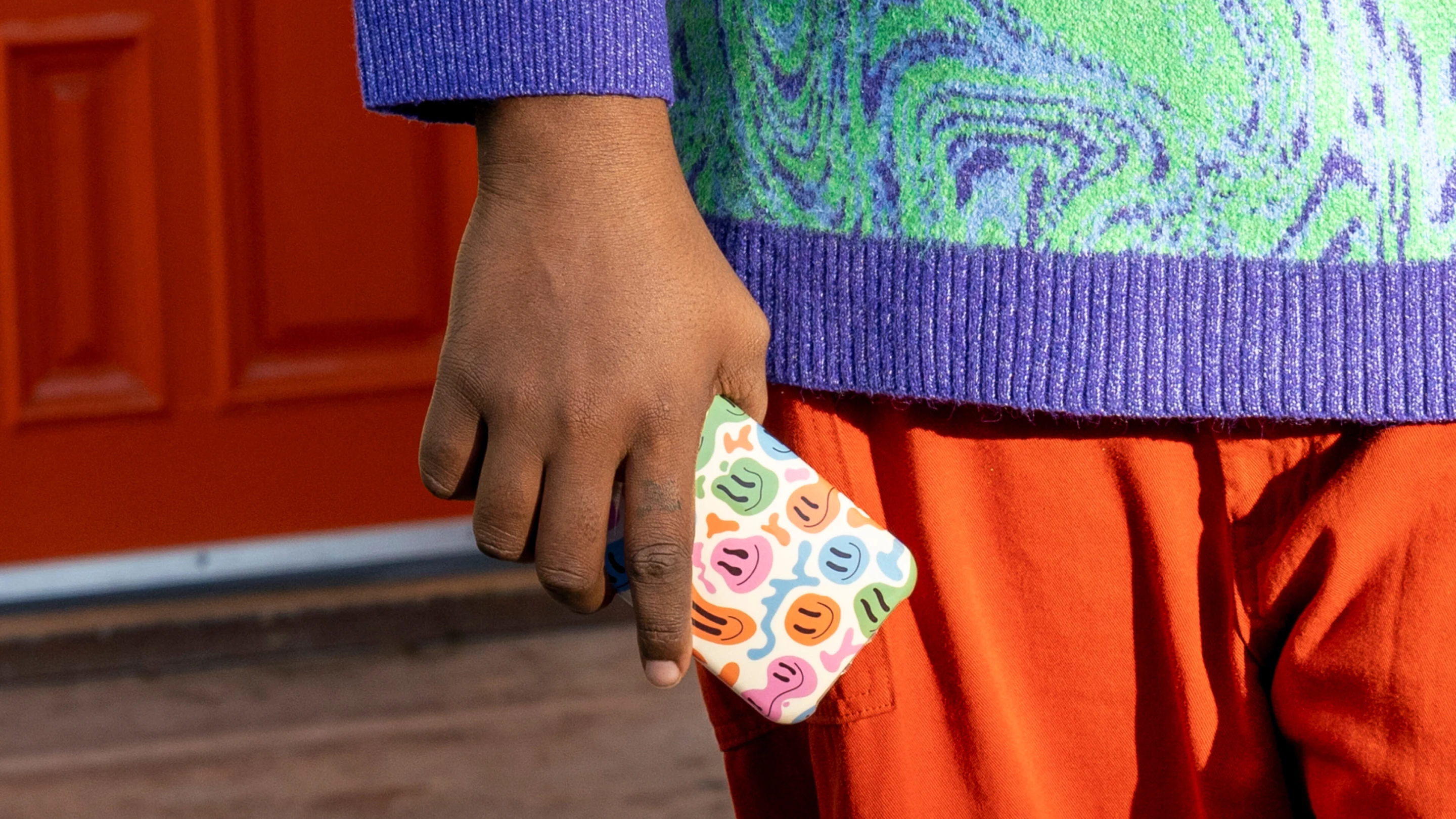 A person wearing a colorful sweater holds a phone in their hand. The phone case is covered in rainbow smiley faces.