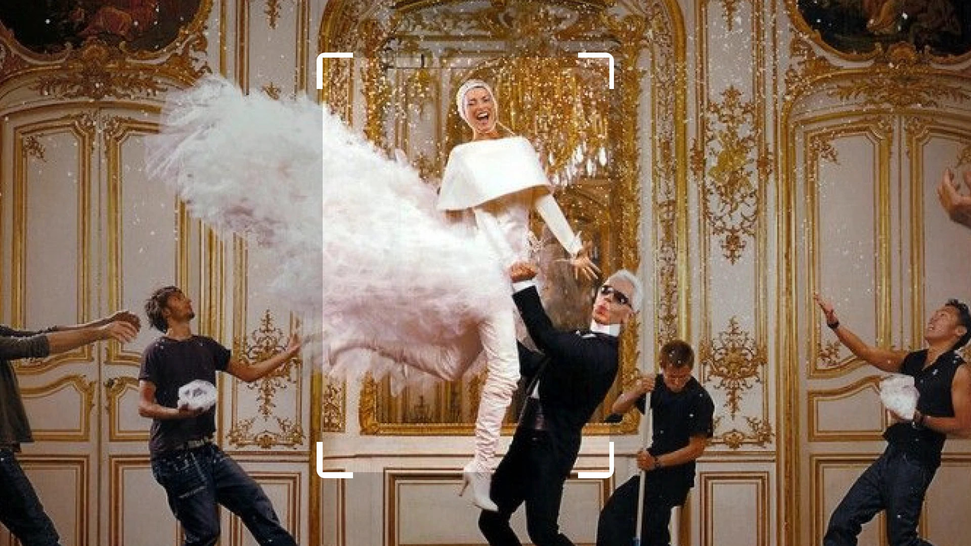 In an ornate golden room, a woman wearing a white outfit adorned with feathers is held up by a man in a black suit