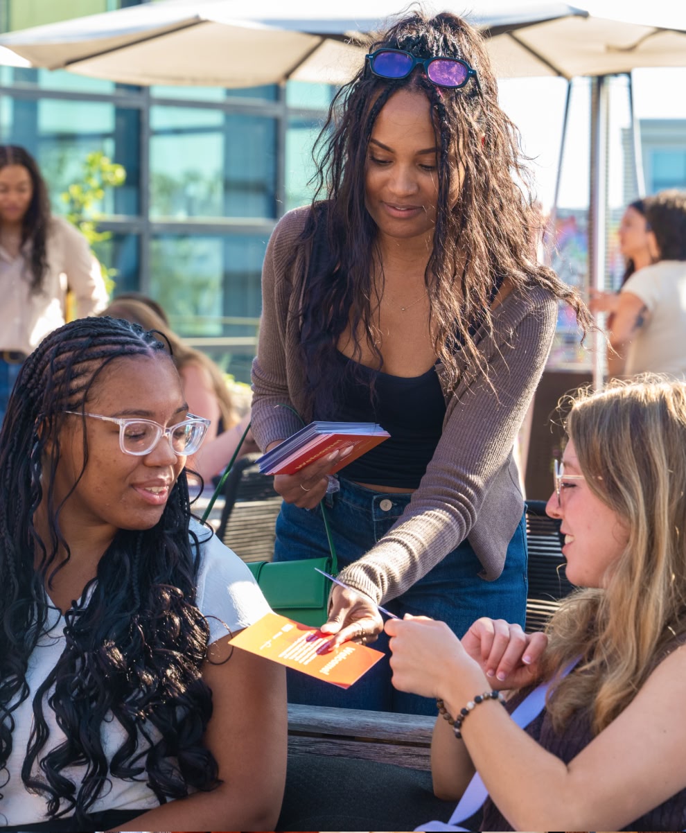 A small group sits outdoors together, smiling and looking at printed cards during a workshop. 