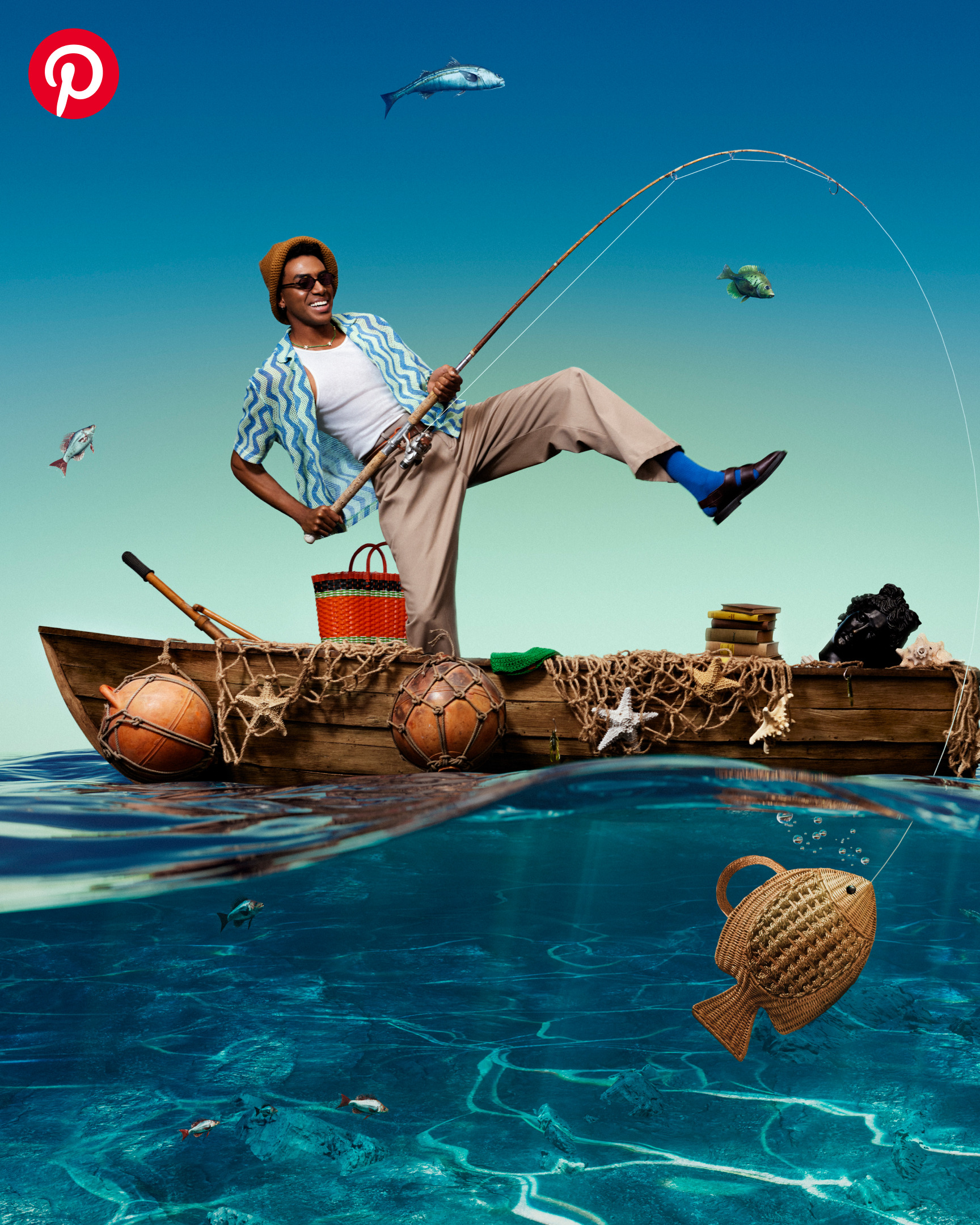A man in khaki pants and a short sleeve button-down stands on one leg in a wooden boat. The boat is draped with fishing nets, and has a wicker goldfish-shaped clutch underneath it. 