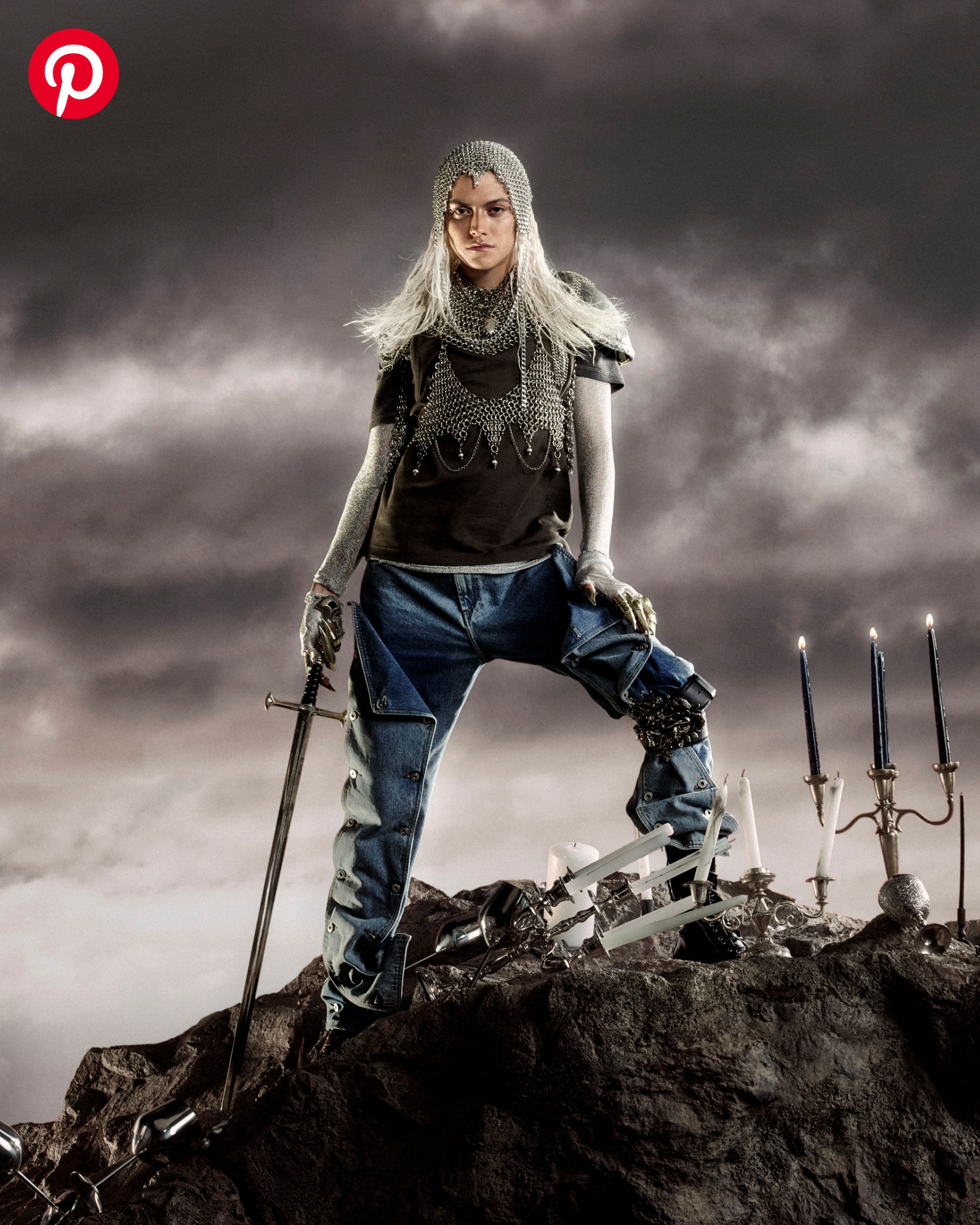 A man wearing a chainmail headpiece and holding a sword poses against a stormy sky surrounded by ornate candelabras