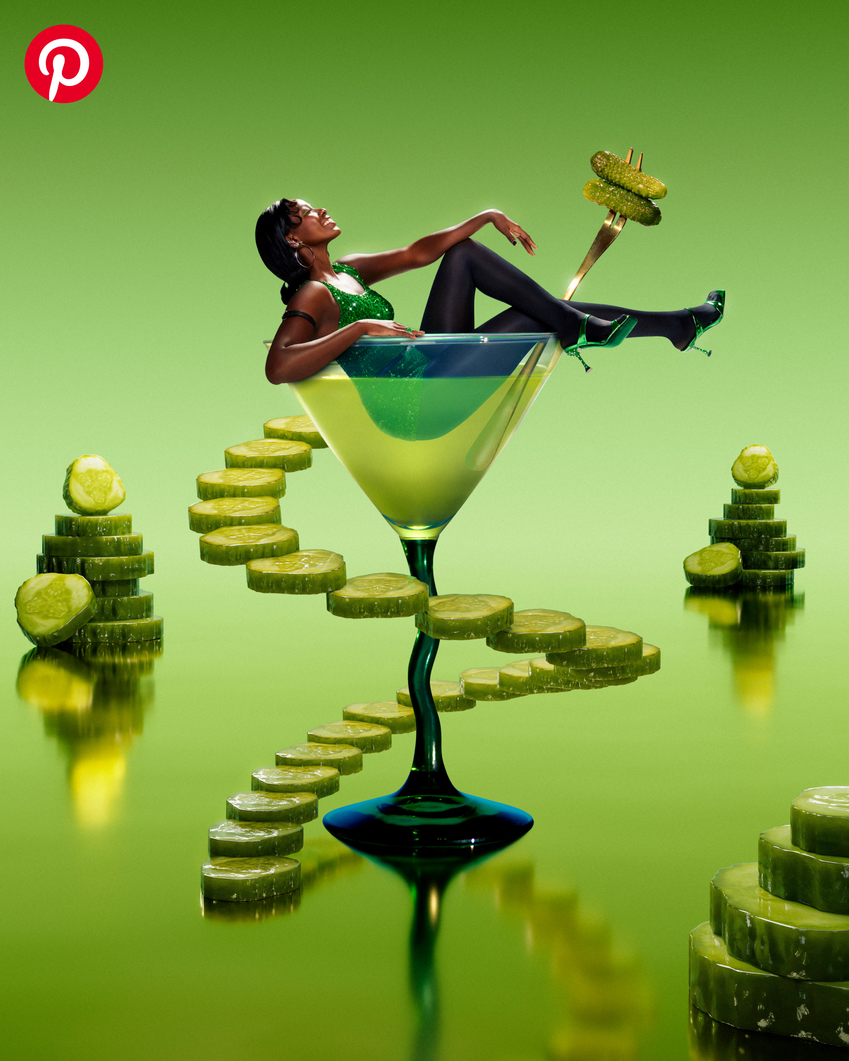 A woman in a green sparkly dress, black tights and metallic green strappy heels sits in an emerald green martini glass. The glass has a long dark green stem and sits against a bluish-green background.