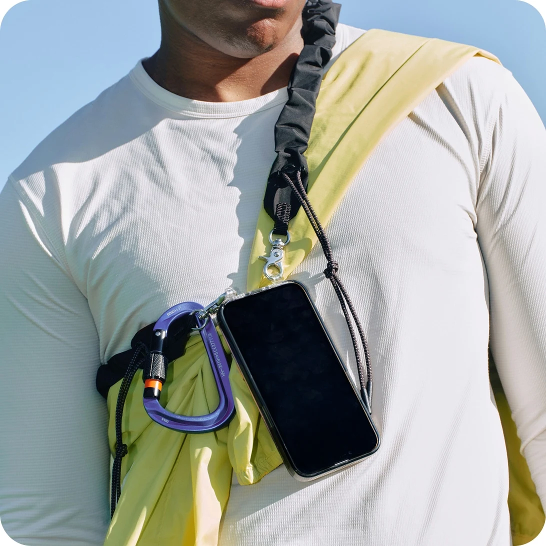 A close-up of a person wearing a crossbody strap with metal clip and a phone case against a blue sky.