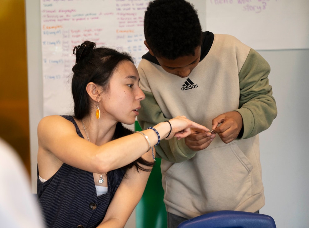 Woman helping a child with a hands-on activity in a classroom setting. They both focus closely on a coin in the child’s hands.