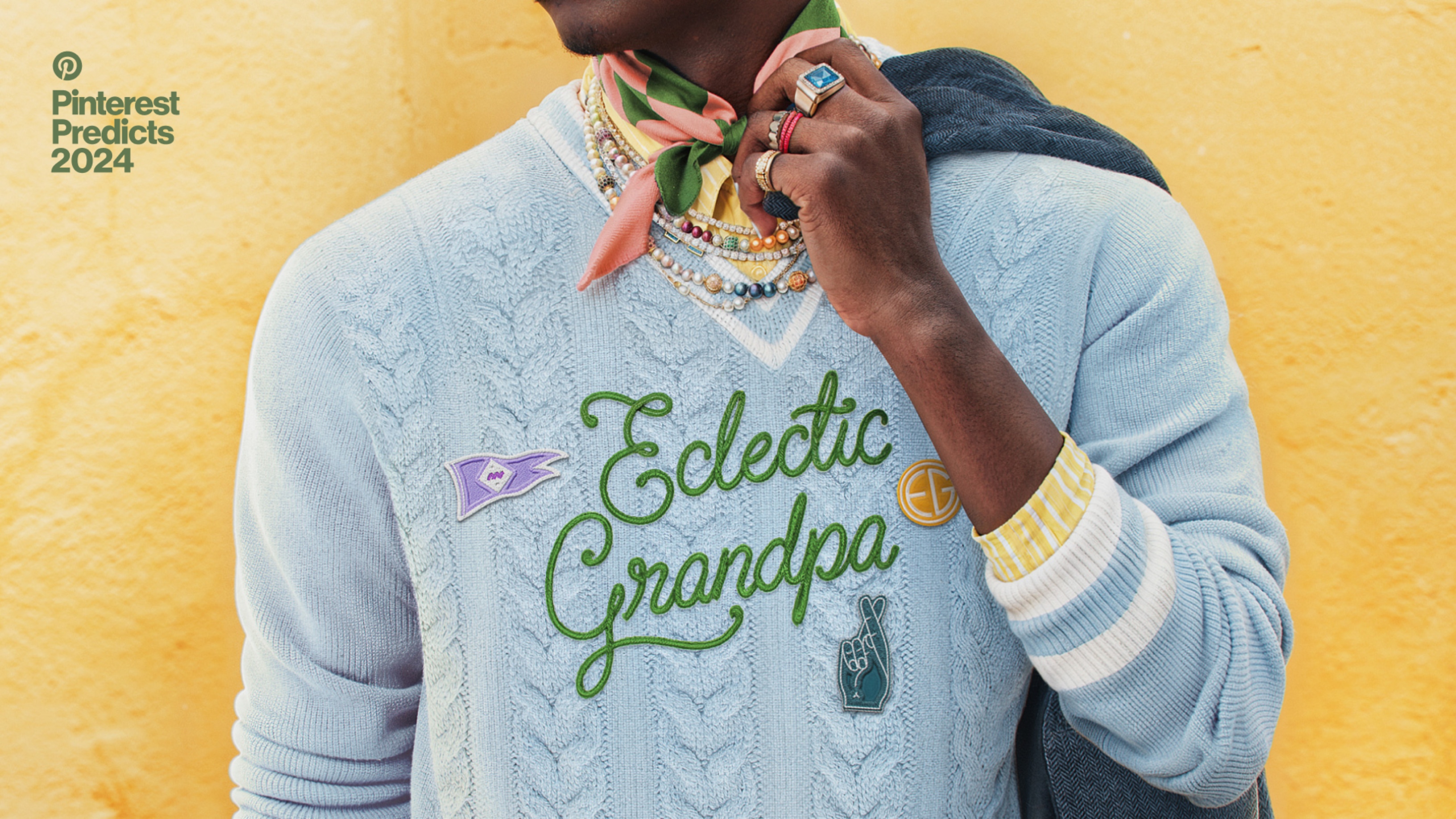 man wearing a blue woven sweater with a yellow striped shirt underneath, styled with bead necklaces, a green and pink scarf, and rings