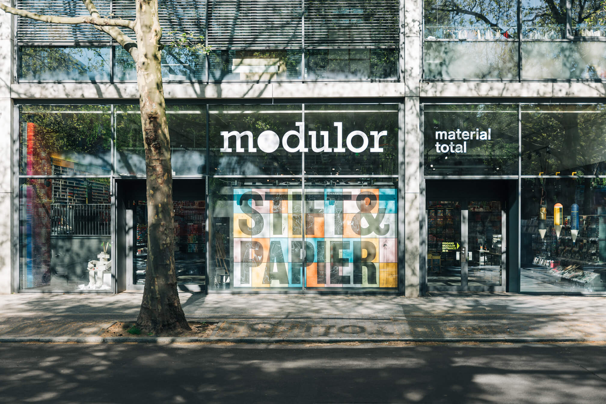 Außenansicht des Modulor-Ladens in Berlin. Die große Glasfassade zeigt die „Stift & Papier"-Kampagne auf bunten Panels.
