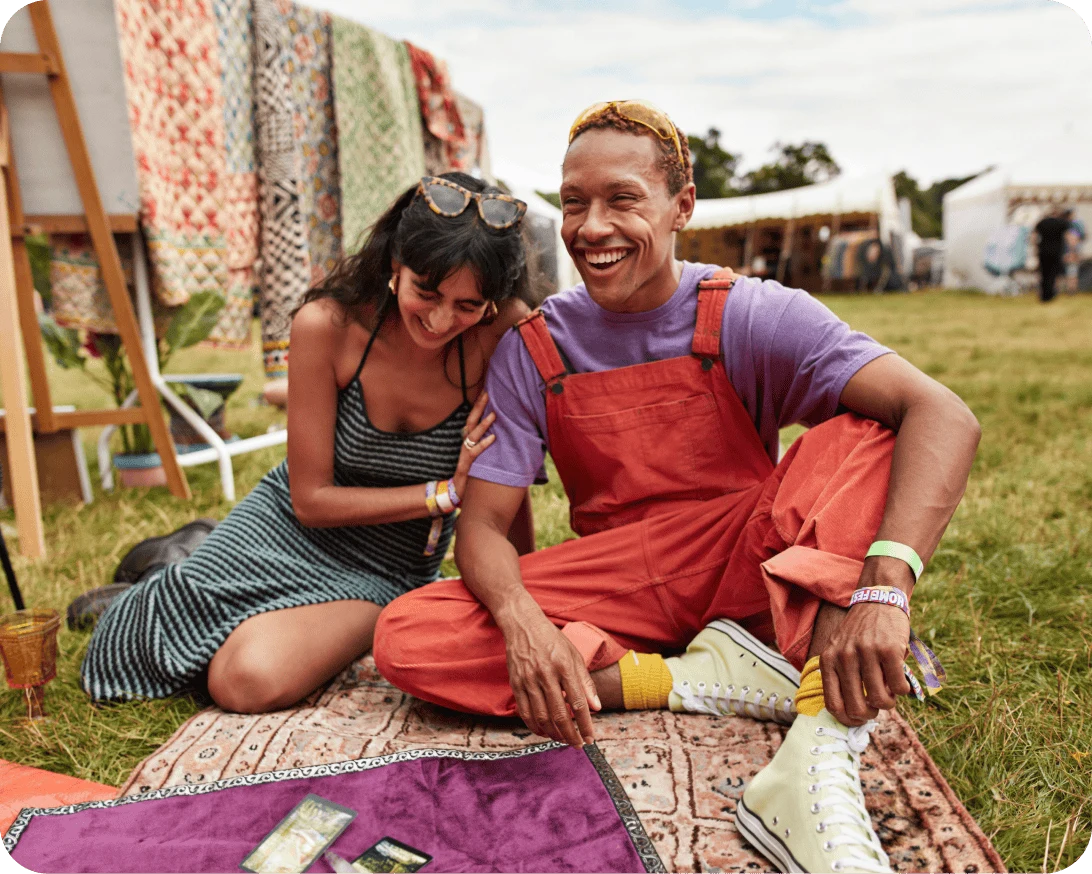 Two smiling people sit on a blanket at an outdoor market.