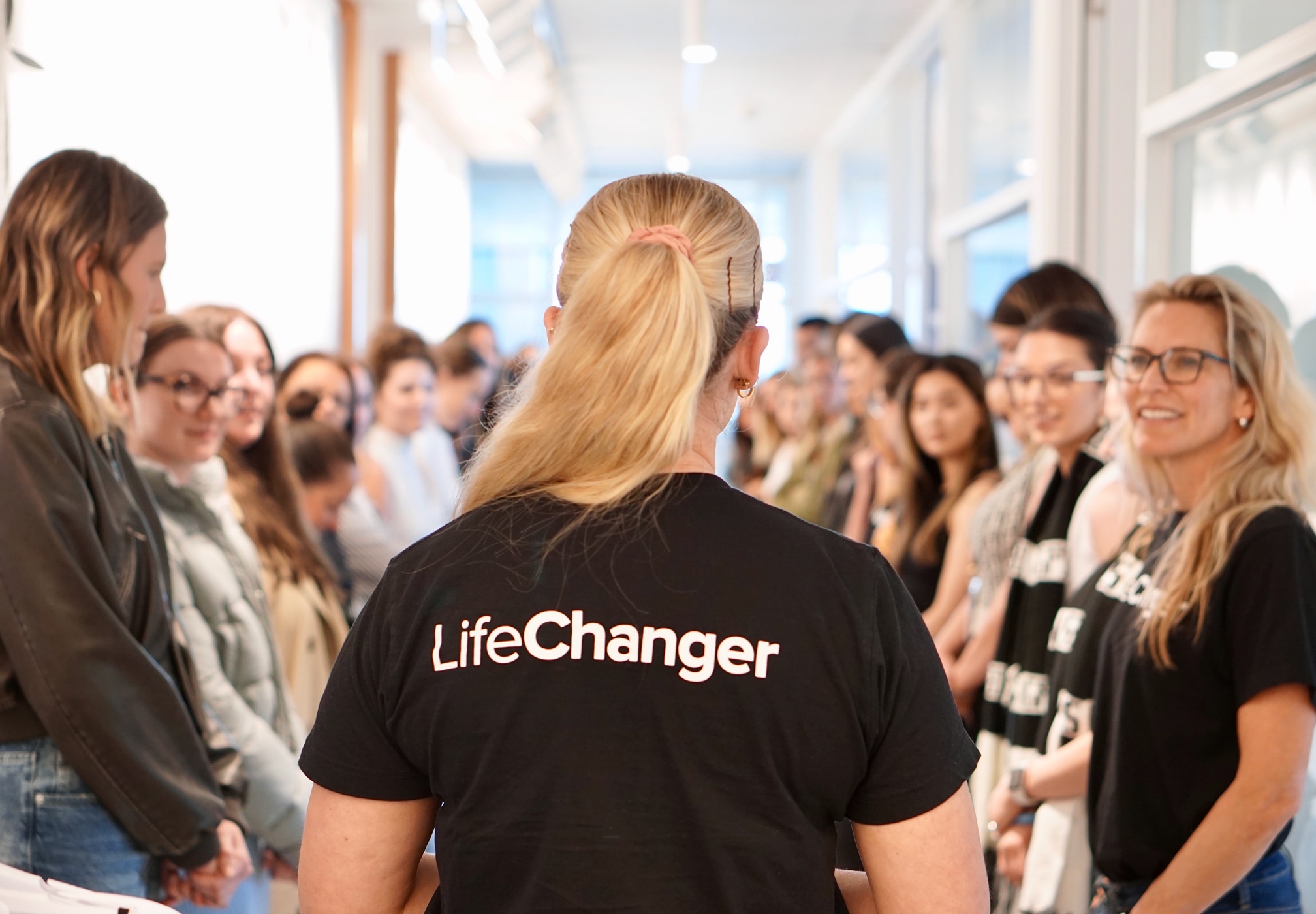 The back of a person wearing a black shirt with text that reads, “LifeChanger” in the middle of a hallway addressing two rows of smiling people lined up on either side.