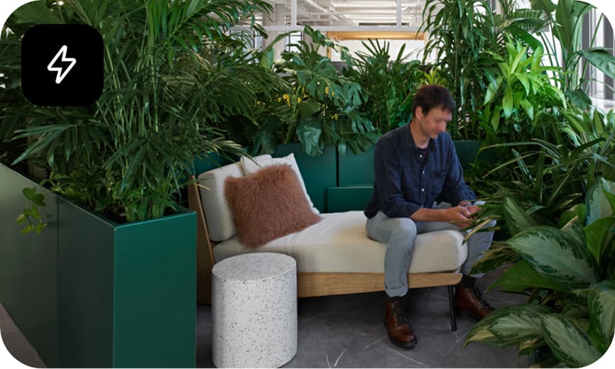 A man in an office sits on a white couch looking on a phone. He is surrounded by plants.