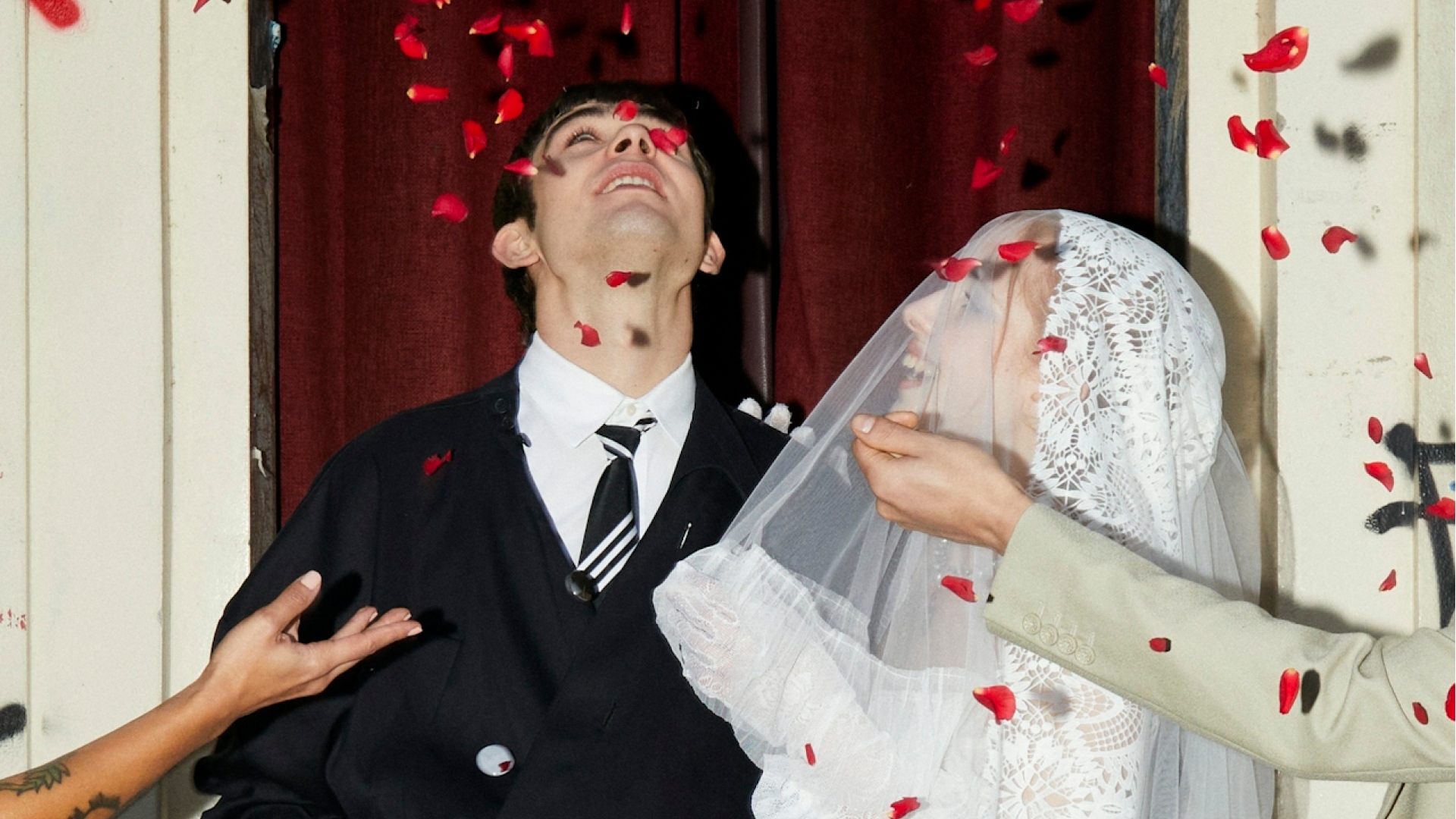 Pareja sonriendo bajo una lluvia de confeti de pétalos rojos; la novia lleva velo y guantes de encaje
