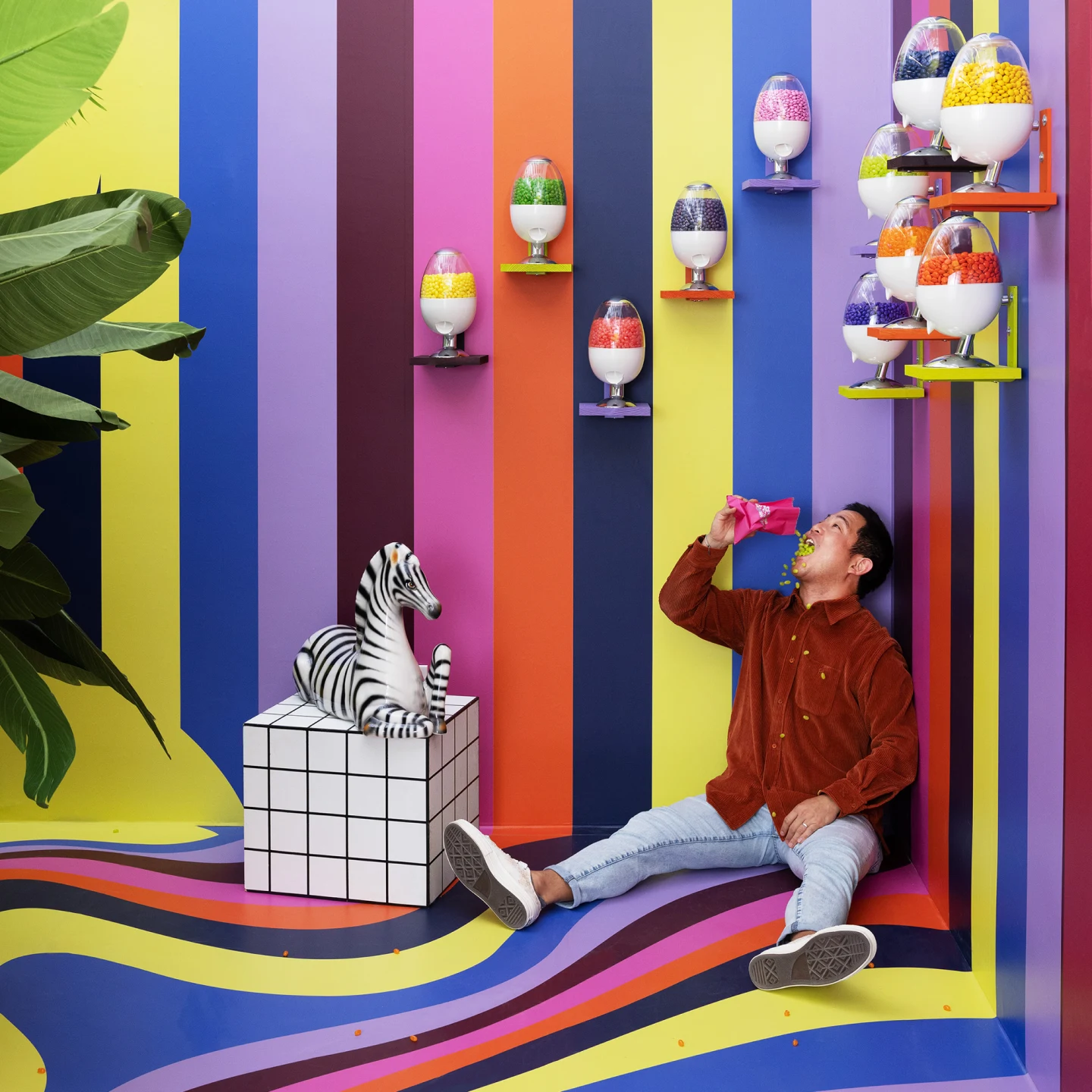  man sits on the floor in a colorful striped room, pouring jellybeans into his mouth.