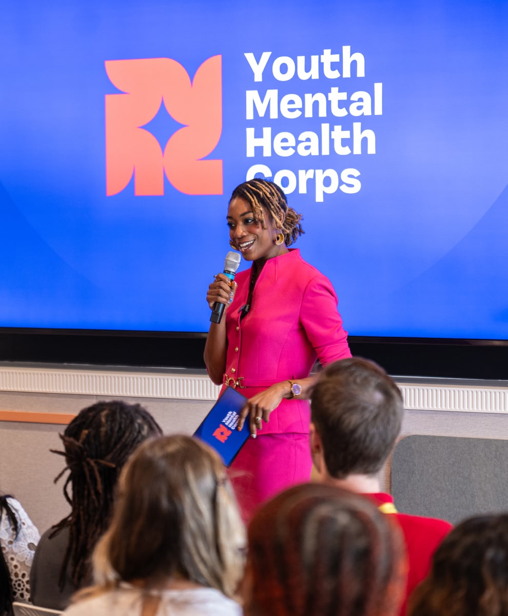 A person speaks into a microphone in front of a large screen that displays “Youth Mental Health Corps,” addressing a seated audience. 