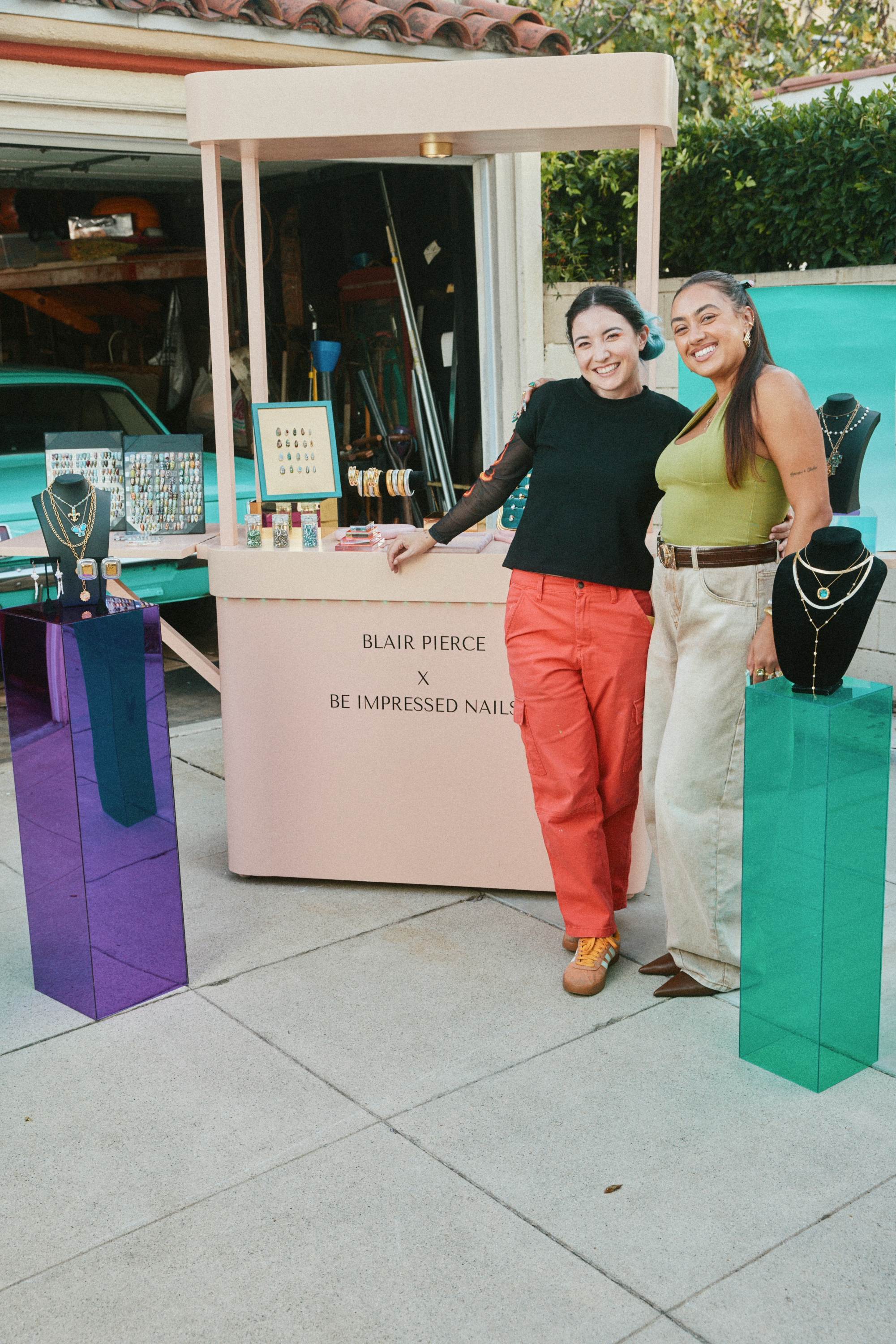 Two women smile and pose beside a small outdoor jewellery pop-up stall, with necklaces and earrings displayed on colourful stands.