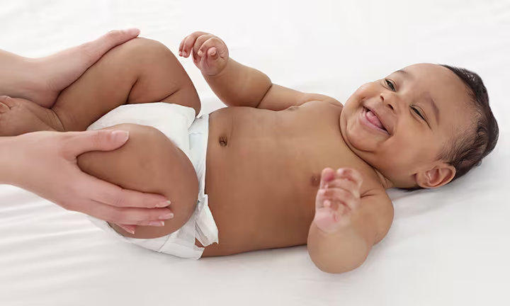 Lächelndes Baby in Windel liegt auf dem Rücken, während eine erwachsene Person sanft seine Beine für eine Massage hält.]