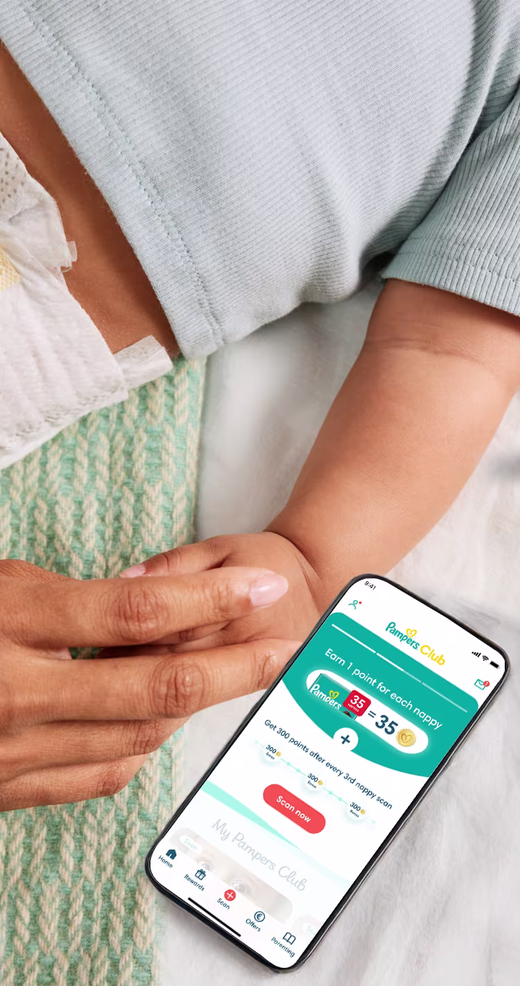 The image shows a close-up of a baby wearing a diaper and a light blue top, with an adult hand gently holding the baby's hand. Beside them, a smartphone displays the "Pampers Club" app, showing a loyalty program to "Earn 1 point for each nappy" and bonus points for scanning every third nappy.