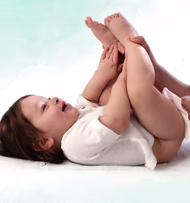 Um bebé está deitado de costas sobre uma superfície branca, levantando os pés enquanto um pai utiliza uma toalhita para limpar o rabo do bebé. Um bebé está deitado de costas sobre uma superfície branca, levantando os pés enquanto um pai utiliza uma toalhita para limpar o rabo do bebé.