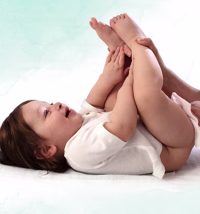 Um bebé está deitado de costas sobre uma superfície branca, levantando os pés enquanto um pai utiliza uma toalhita para limpar o rabo do bebé.