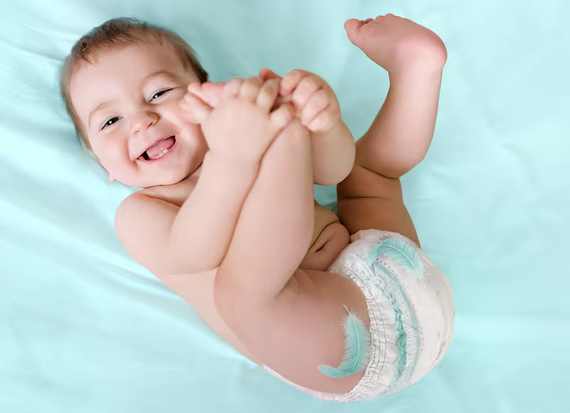 Un bebé feliz tumbado de espaldas sobre una sábana azul claro, sosteniendo sus pies y llevando un pañal-braguita  Dodot.