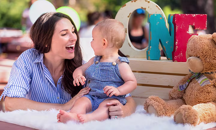 Smiling woman and baby in matching blue outfits celebrating first birthday with a teddy bear and colorful "ONE" sign.