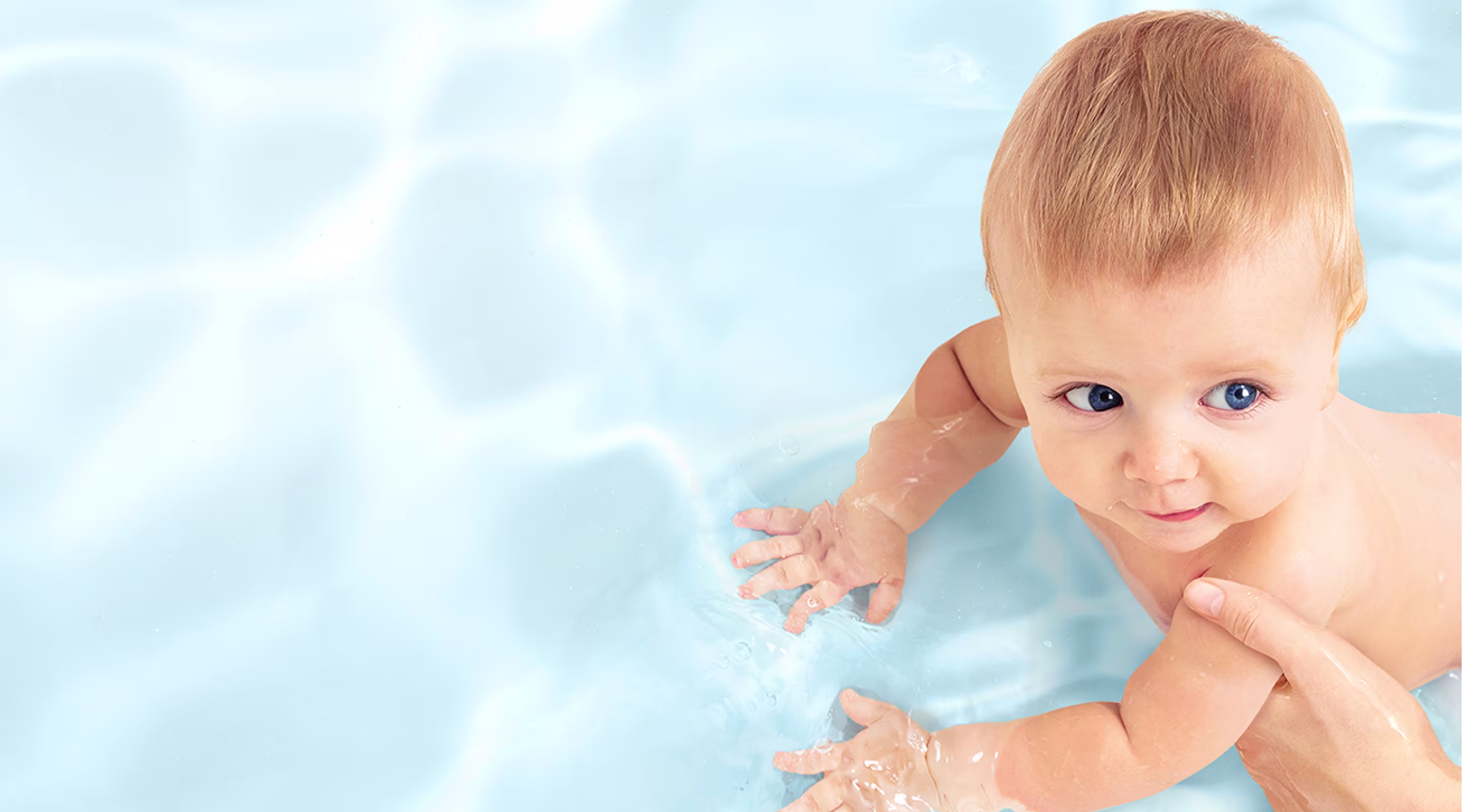 Eine Draufsicht auf ein Baby, das sanft von der Hand eines Erwachsenen in klarem, blauem Wasser gestützt wird.