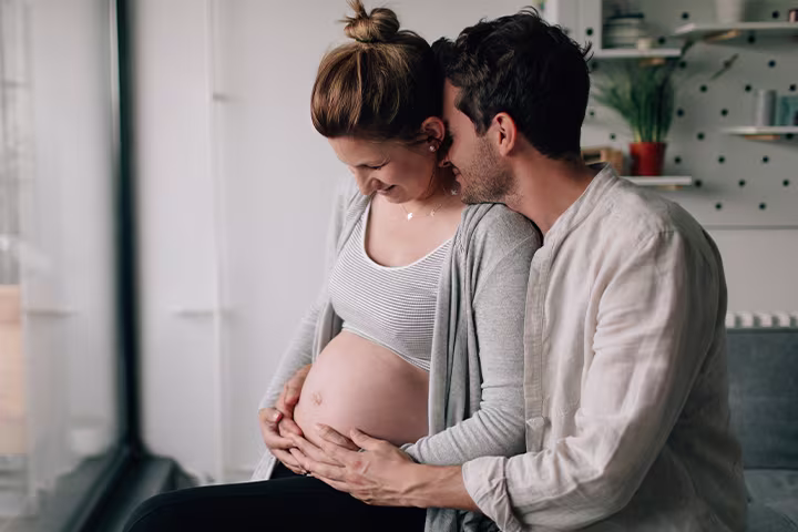 Cuplu care se îmbrățișează, în timp ce bărbatul mângâie burtica  femeii însărcinate.

