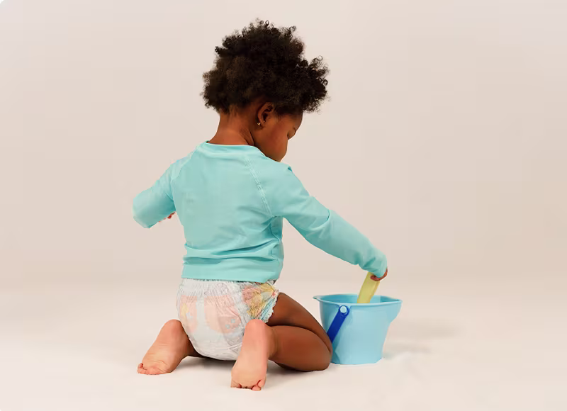 Uma criança pequena que usa Dodot Splashers e uma t-shirt de banho cor teal ajoelha-se enquanto brinca com uma pá e um balde.