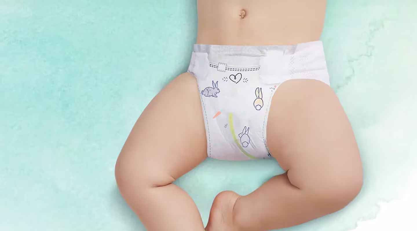 En babys nedre torso och ben visas från en ovanifrån vy, liggande på en ljusgrön yta. Pampers-blöjan framhäver en fuktindikator.