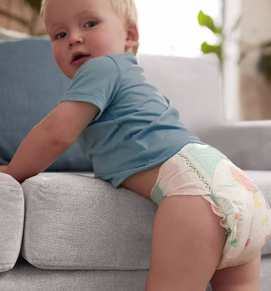 Um bebé olha por cima do seu ombro direito com uma expressão curiosa, usando uma fralda da Pampers.