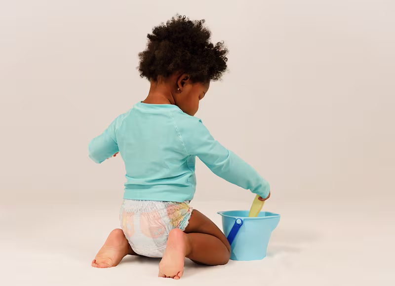 Un băiețel mic în scutece Pampers Splashers și tricou de baie turcoaz stă în genunchi, jucându-se cu o lopățică și o găletușă. Un băiețel mic în scutece Pampers Splashers și tricou de baie turcoaz stă în genunchi, jucându-se cu o lopățică și o găletușă.