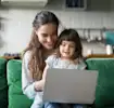 Smiling mother and child using a laptop together on a green couch in a cozy living room.