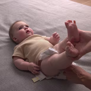 A baby lying on their back during a diaper change. A parents hand is lifting the babies feet up to pull the used diaper out. The baby is dressed in a yellow t-shirt. all sensitive parts are covered.