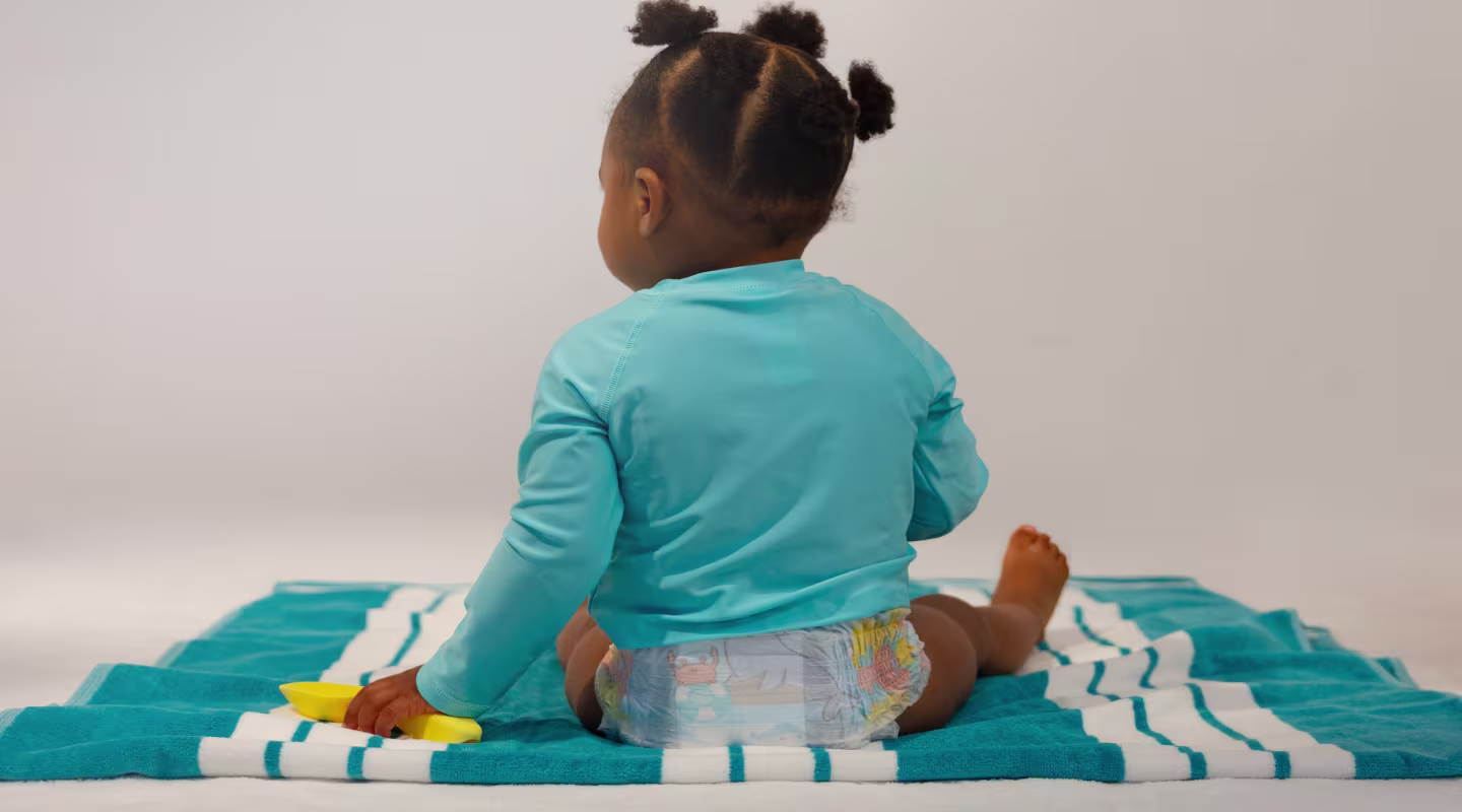 A cheerful baby sits on a vibrant blue towel, showcasing their Pampers Splashers from behind, ready for a fun day at the beach!