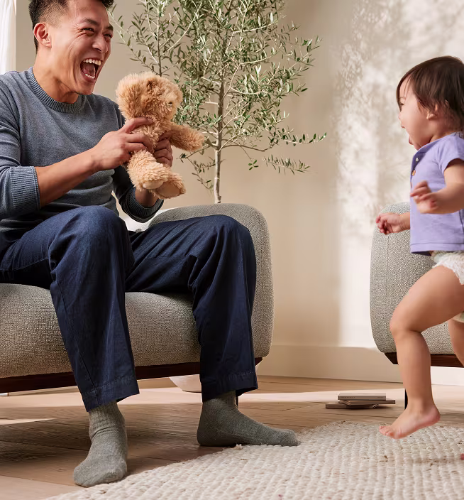Baby and dad happily playing next to the sofa with a plush teddy. Both are laughing and smiling