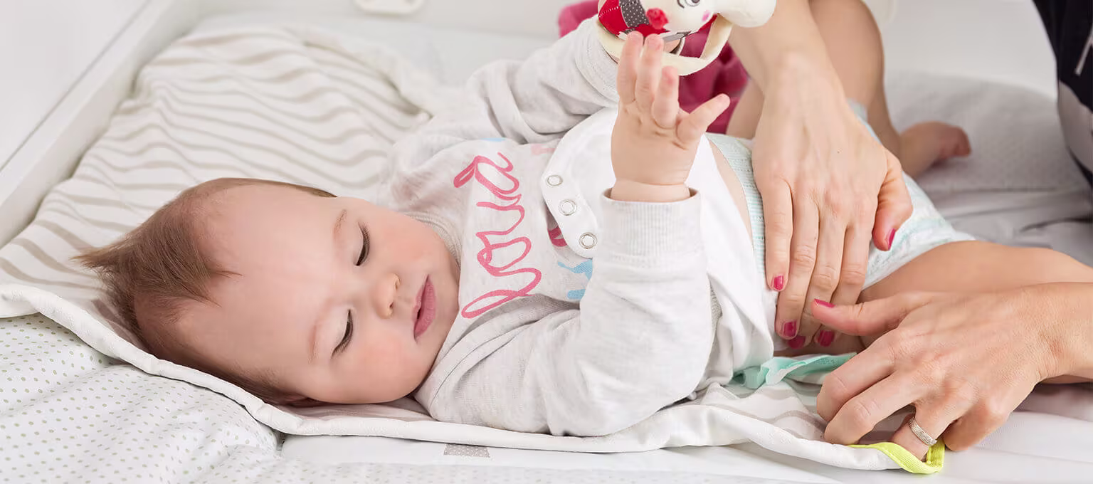 An infant lying on a changing mat, holding a toy as a caregiver changes their diaper