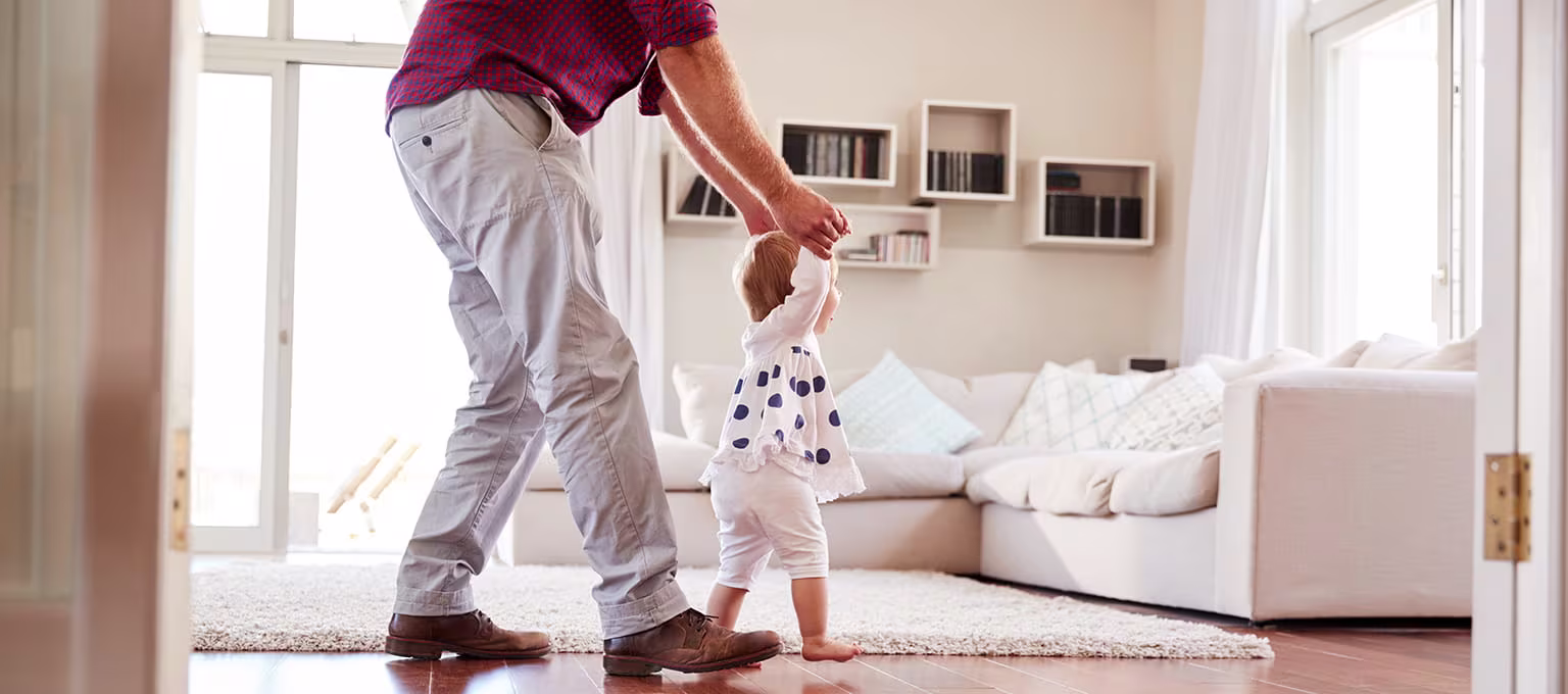 Père aidant son tout-petit à marcher dans un salon lumineux, tenant ses mains au-dessus du sol en bois.