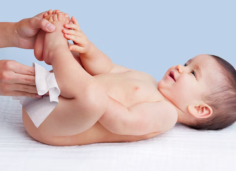 Un bebé yace de espaldas sobre un cambiador blanco con las rodillas dobladas, mirando hacia arriba mientras las manos de una persona le limpian suavemente el trasero con una toallita blanca. Un bebé yace de espaldas sobre un cambiador blanco con las rodillas dobladas, mirando hacia arriba mientras las manos de una persona le limpian suavemente el trasero con una toallita blanca.