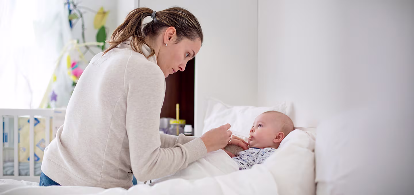 Foto einer Mutter, die ihr Baby im Bett mit einem Löffel füttert, während sie sich über das Kind beugt und es aufmerksam ansieht.